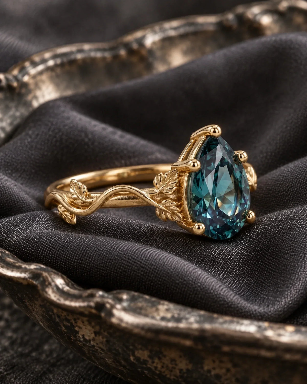 Gold pear-shaped teal gemstone ring with carved leaf shoulders resting at a side angle on charcoal fabric in an antique silver dish