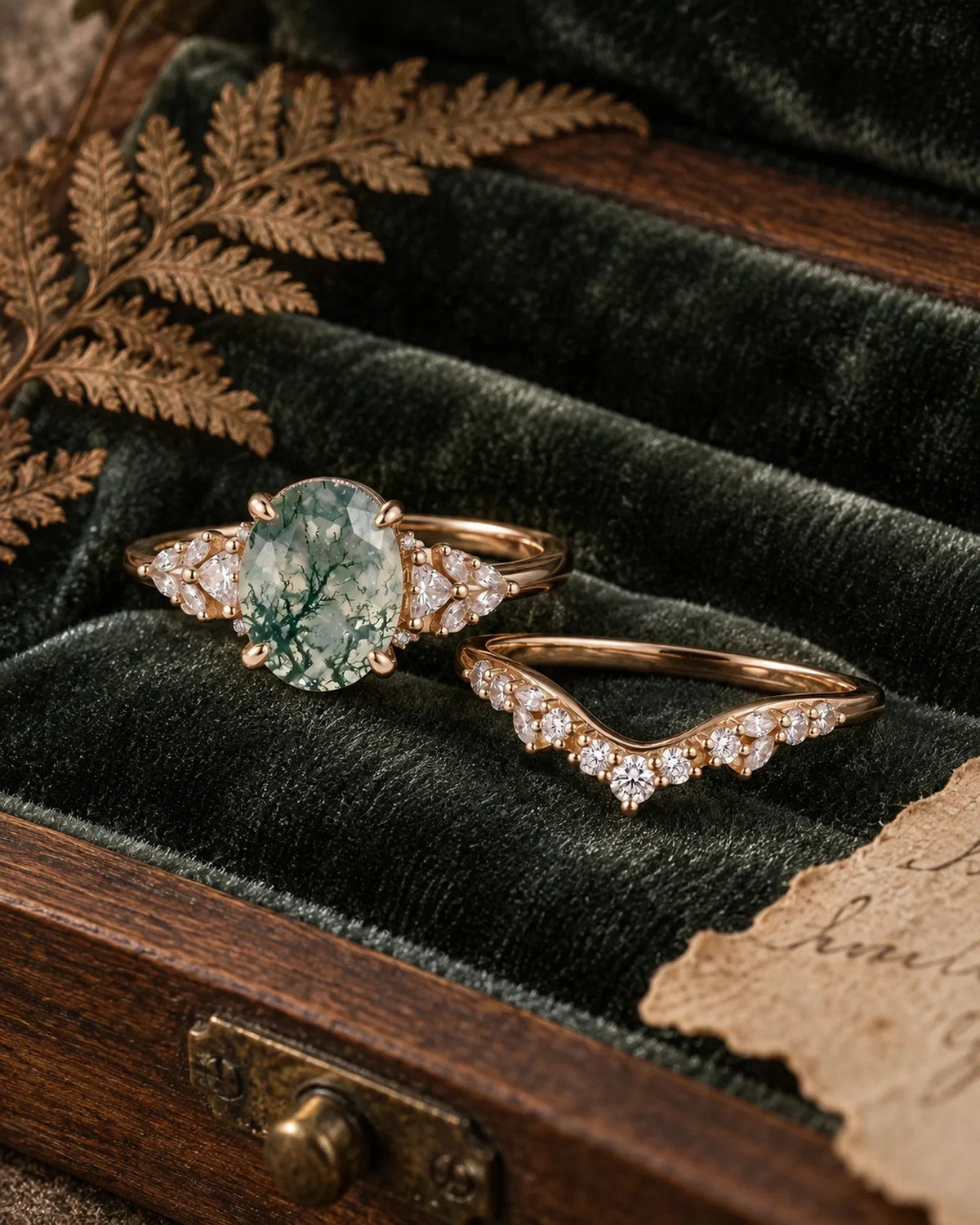Oval moss agate bridal ring set arranged at an angle on dark green velvet with a dried fern and antique wood details
