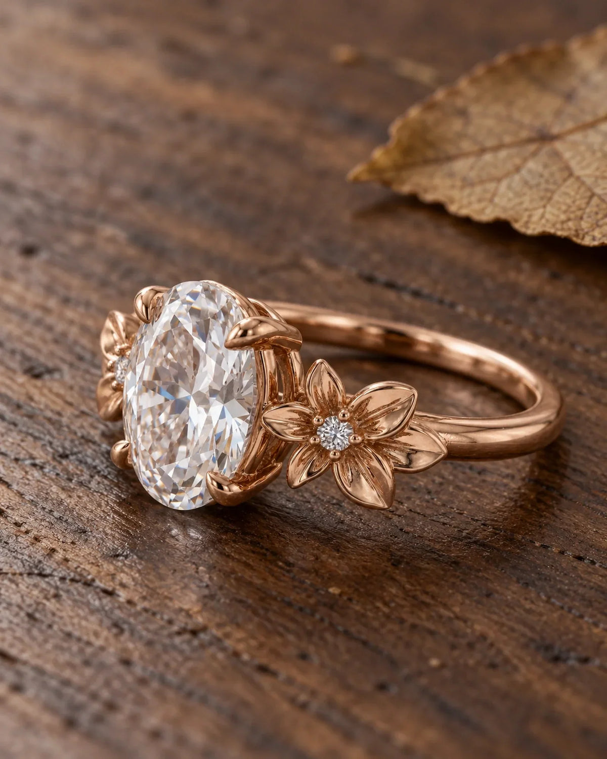 Rose-gold floral engagement ring resting diagonally on dark walnut beside a dry leaf, with its raised center setting visible