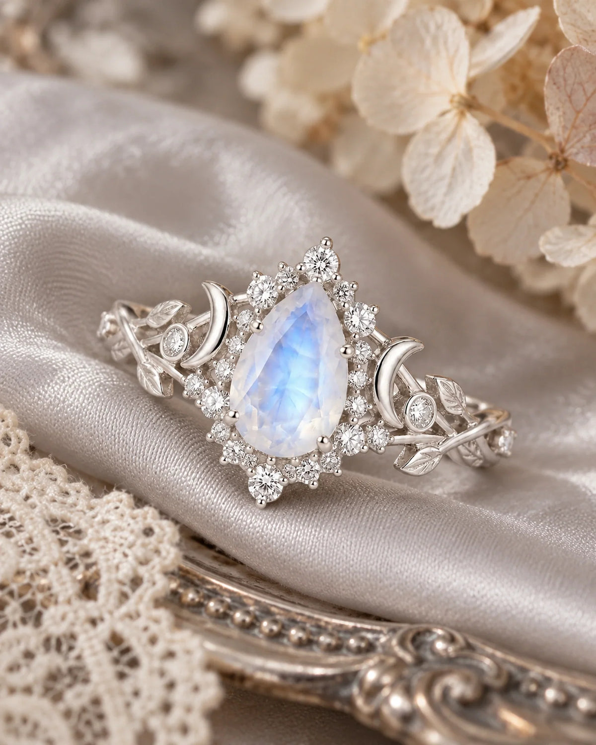 Lunavelle pear moonstone halo ring with sculpted crescent moons and engraved leaf shoulders resting diagonally on silk beside antique lace