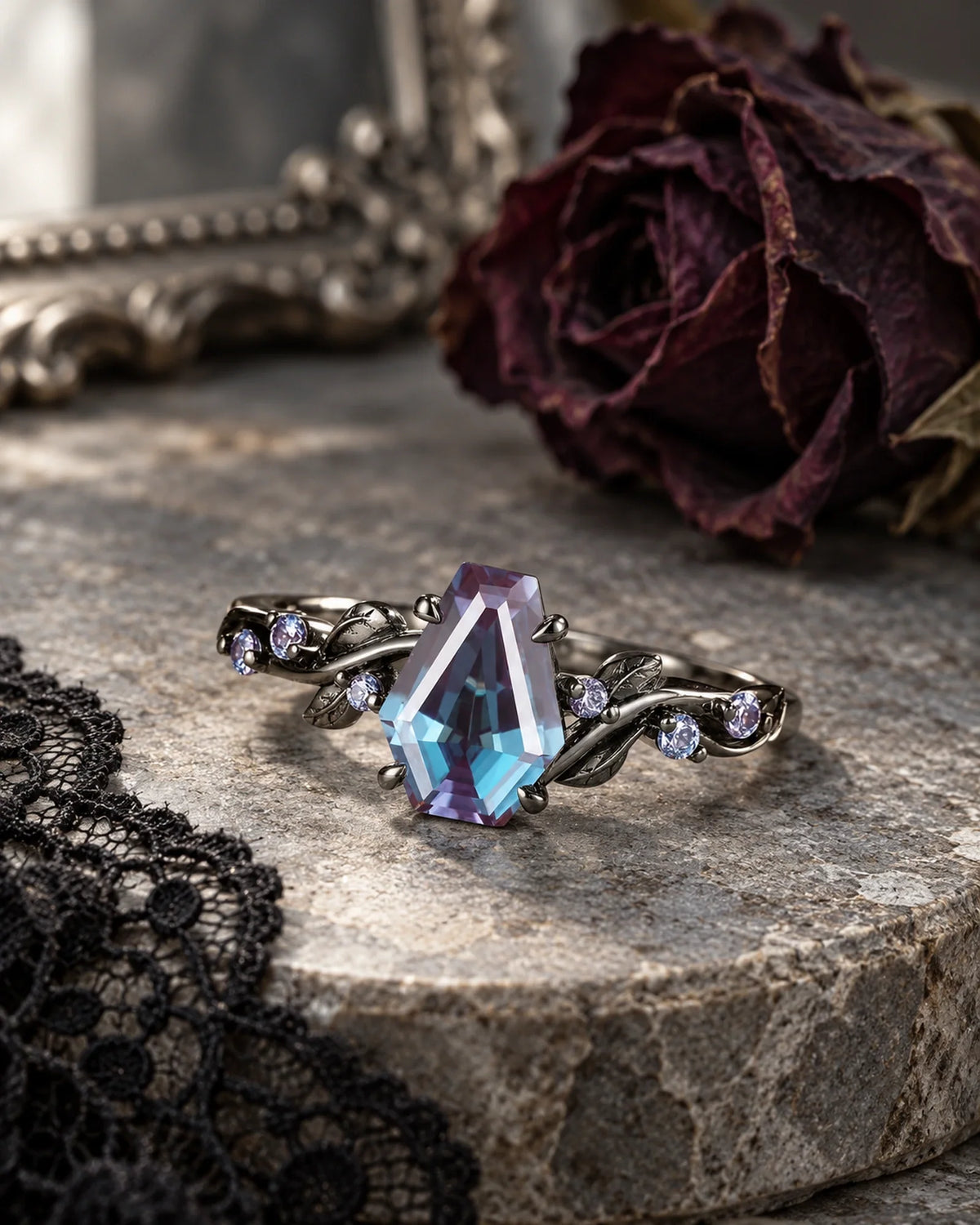 Shadowleaf coffin-shaped blue-violet gemstone ring with dark leaf shoulders on aged gray stone beside black lace and a dried rose
