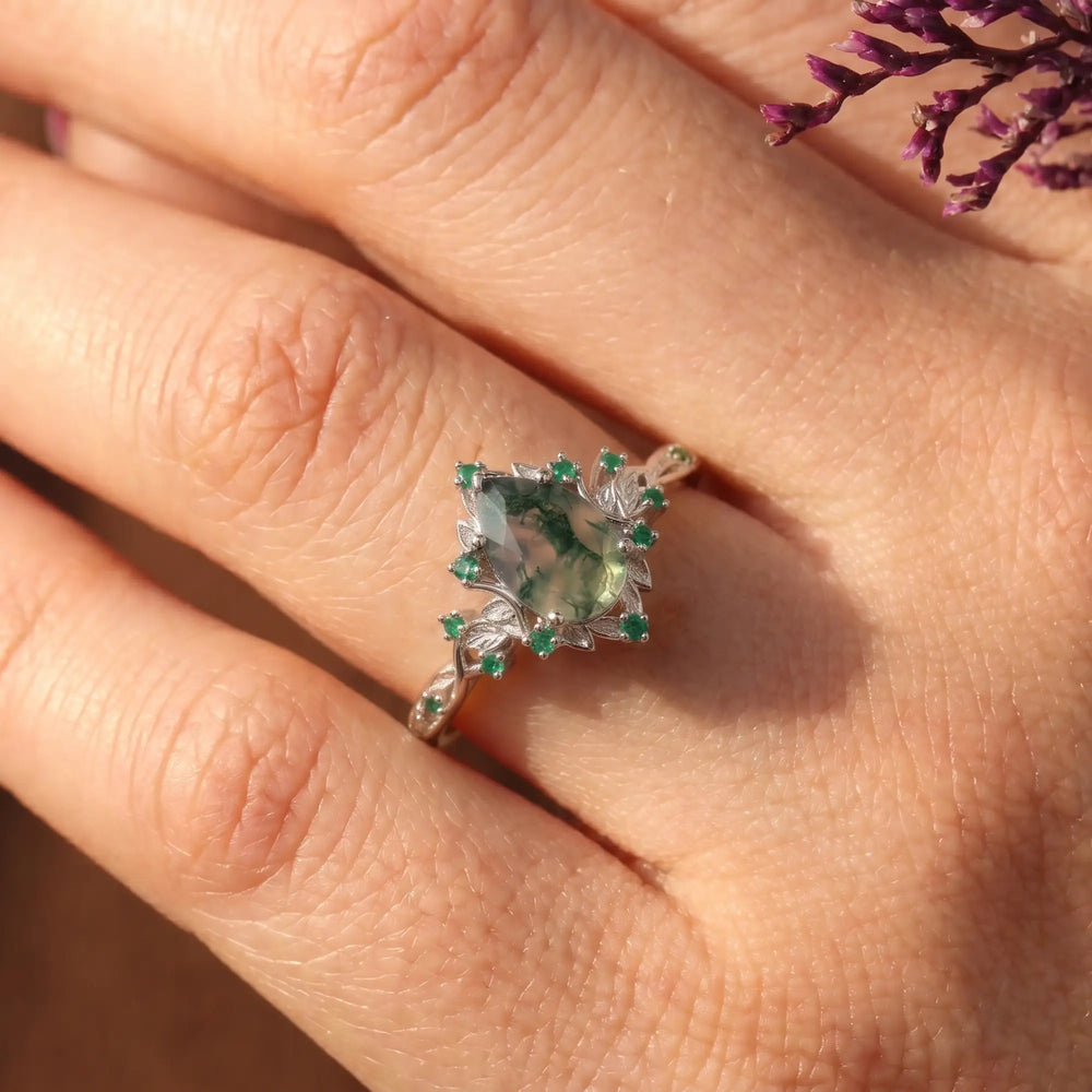 Lunavelle pear cut moss agate engagement ring worn on a finger, displaying its forest-like gemstone inclusions, delicate botanical band and crescent moon design.