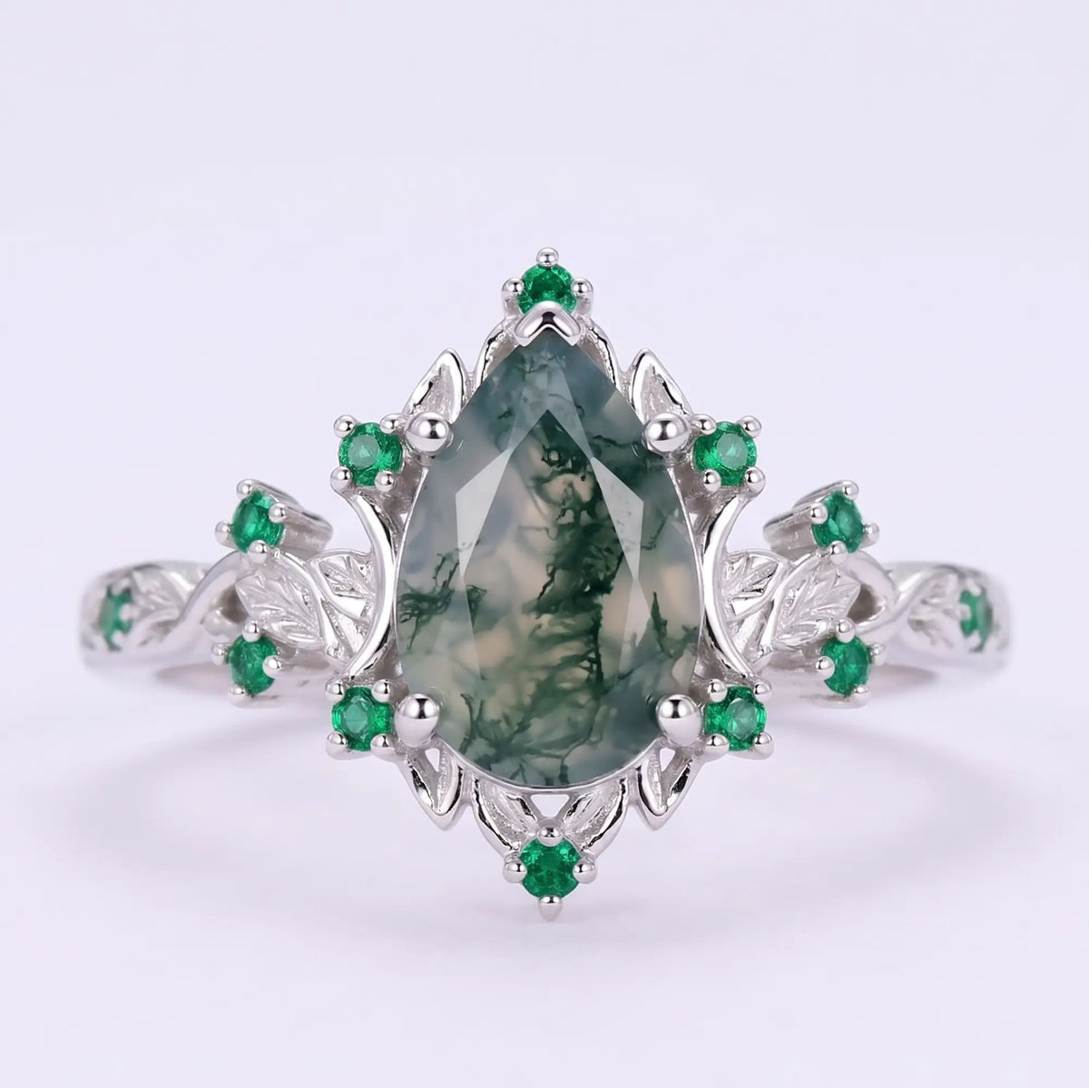 Front view of Lunavelle pear cut moss agate engagement ring with natural emerald cluster accents, crescent moon motifs and botanical leaf details in white gold.